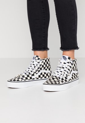 Stopy w czarno-białych wysokich trampkach Vans w kratkę, z białymi sznurowadłami, ubranych w czarne podwinięte dżinsy z postrzępionymi brzegami.