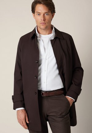 Brązowy, lekki trencz z kołnierzem, z przodu zapinany na guziki, z długimi rękawami. Nosi się go na białej koszuli i brązowych spodniach.