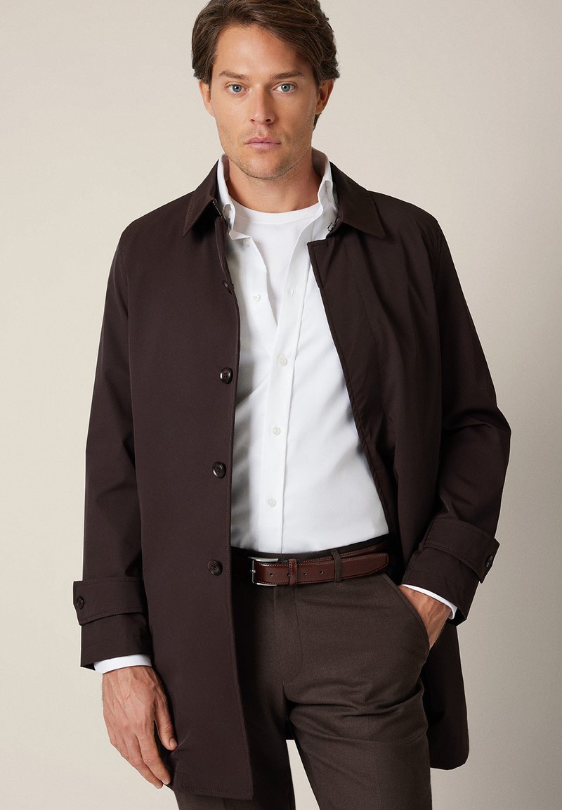 Brązowy, lekki trencz z kołnierzem, z przodu zapinany na guziki, z długimi rękawami. Nosi się go na białej koszuli i brązowych spodniach.