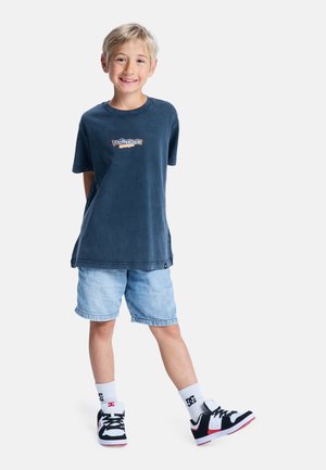 Enfant souriant portant un t-shirt graphique bleu marine, un short en jean clair, des chaussettes blanches et des baskets noir et blanc, debout les mains dans le dos.