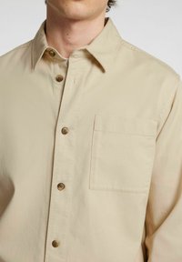 Camicia beige con bottoni, realizzata in cotone, con colletto classico, taschino sul petto e bottoni marroni davanti. Texture liscia.