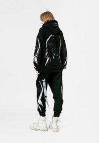 Sudadera con capucha negra y pantalones deportivos con diseños de alas bordados; presenta acentos blancos y metálicos, tejido texturizado y un detalle de etiqueta. Botas blancas.