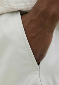Pantalon beige clair avec un détail en tissu blanc texturé à la taille, révélant un teint de peau marron en dessous. Surface lisse avec des coutures soignées.