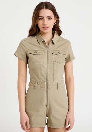 Młoda kobieta ubrana w beżowy kombinezon z krótkim rękawem i suwakiem z przodu, z kieszeniami na piersiach i kołnierzykiem, stojąca na tle jednolitego białego tła.