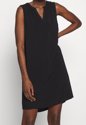 Robe noire sans manches avec décolleté en V portée par une personne avec un tatouage sur l'avant-bras gauche et un collier en chaîne dorée, posant avec la main gauche sur la hanche.
