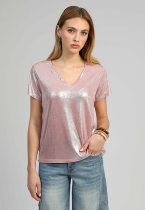 Jeune femme aux longs cheveux blonds portant un haut rose brillant à col en V, un jean bleu et plusieurs bracelets au poignet droit, debout devant un fond gris.