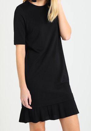 Kobieta ubrana w czarną sukienkę z krótkim rękawem i plisowanym brzegiem, stojąca z jedną ręką przy twarzy na tle jasnego, jednolitego tła.