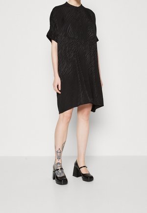 Schwarzes Kurzarmkleid mit strukturiertem, plissiertem Design, ausgestattet mit hohem Kragen und Knopfverschluss. Getragen mit glänzenden, schwarzen Blockabsatzschuhen.