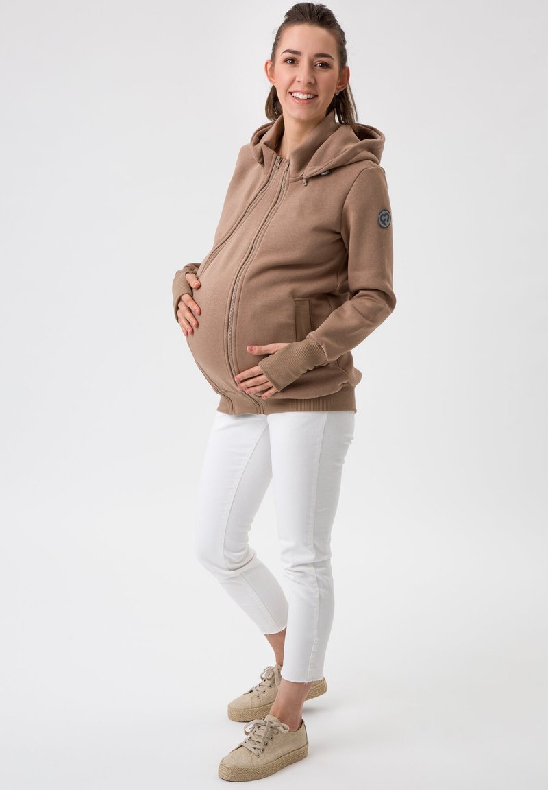 Veste à zip de maternité en tissu marron clair, dotée d'une poche kangourou et d'ouvertures pour les pouces dans les manches. Associée à un pantalon blanc ajusté et des chaussures légères.