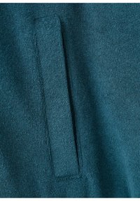 Gros plan sur un tissu bleu sarcelle avec une poche passepoilée rectangulaire verticale cousue à la surface du vêtement.