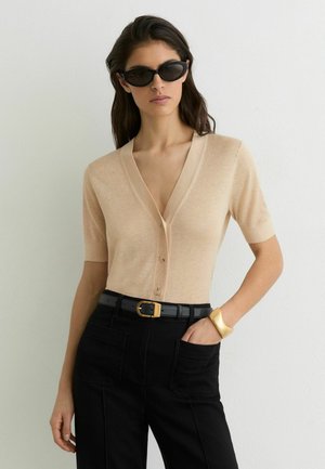 Frau trägt beiges kurzärmeliges V-Ausschnitt-Oberteil mit Knöpfen, schwarze Hose mit Gürtel, Katzenaugen-Sonnenbrille und goldenes Manschettenarmband, steht vor weißer Wand.