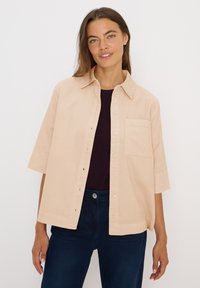 Camisa de manga corta beige con botón en la parte delantera, cuello y bolsillo en el pecho, confeccionada con una tela ligera y con un corte cómodo.
