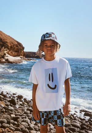 Chłopiec w niebiesko-czarnym kratkowanym kapeluszu typu bucket, w dopasowanych szortach z uśmiechniętymi buźkami i białej koszulce, stoi na kamienistej plaży nad morzem.