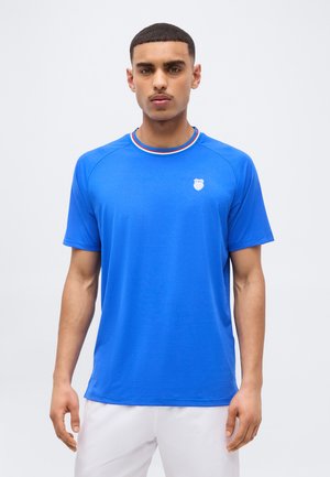 Άνδρας που φοράει φωτεινό μπλε κοντομάνικο μπλουζάκι με μικρό λευκό λογότυπο στο στήθος και κόκκινο, λευκό και μπλε ριγωτό γιακά.