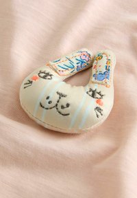 Giocattolo morbido in tessuto a forma di gatto, di colore beige chiaro con strisce blu e dettagli del viso ricamati, su sfondo rosa.