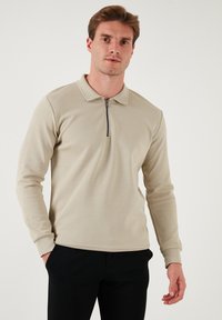 Polo a maniche lunghe di colore beige chiaro con colletto con zip, tessuto texturizzato e design aderente, abbinato a pantaloni neri.
