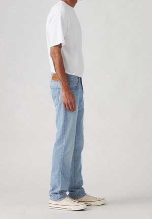 Homme debout de profil portant un t-shirt blanc, un jean bleu clair et des baskets blanches basses sur un fond gris uni.