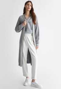 Cardigan lungo grigio con bordo a righe, maglione grigio testurizzato, pantaloni bianchi su misura e sneakers bianche. Il tessuto sembra morbido e accogliente.