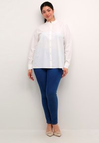 Hvid langærmet skjorte med flæser ved kraven og knaplukning, parret med blå skinny jeans. Let stof med en glat tekstur.