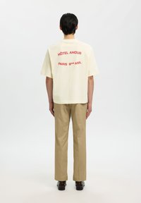 Osoba koja nosi kremastu majicu kratkih rukava s crvenim natpisom "HÔTEL AMOUR PARIS 9ÈME ARR." na leđima, bež hlače i tamne cipele, stojeći.