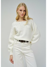 Maglione color crema con scollatura rotonda, impreziosito da piccoli dettagli simili a perle, abbinato a pantaloni bianchi a vita alta e una cintura scura.