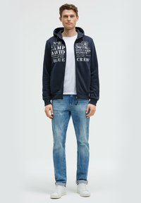 Sudadera con cremallera en color azul marino, con texto impreso, llevada sobre una camiseta blanca, combinada con jeans de un azul claro y zapatillas blancas. Tejido suave y corte casual.