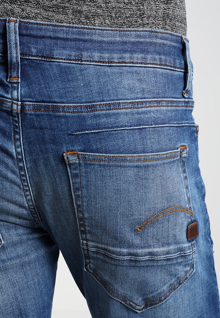 Jeans en denim bleu avec une coupe mi-haute, des coutures visibles et une poche arrière au design distinctif avec une petite étiquette en cuir.