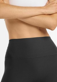 Torso y brazos cruzados de una persona que lleva leggings negros de tiro alto y canalé frente a un fondo blanco liso.