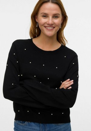 Pull en tricot noir avec un col rond, orné de petites perles. Manches longues et coupe décontractée. Porté avec un jean en denim bleu.