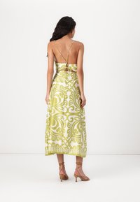 Vestido maxi com um padrão ornamental em verde e branco. Apresenta alças finas, costas abertas e um design fluido. Usado com sandálias de tiras.