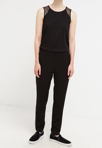Schwarzer Overall mit Spitzenakzenten an den Schultern, ärmelloses Design, schmal zulaufende Beine und kombiniert mit schwarzen Slippern. Glatte Stofftextur.