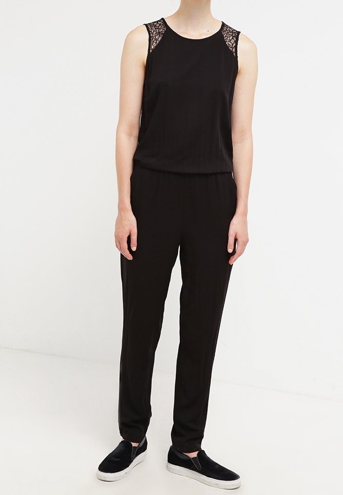 Schwarzer Overall mit Spitzenakzenten an den Schultern, ärmelloses Design, schmal zulaufende Beine und kombiniert mit schwarzen Slippern. Glatte Stofftextur.