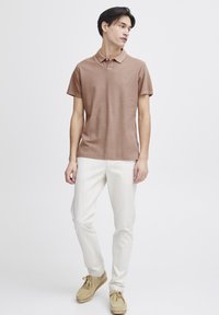 Pruun polo särk, lühikesed varrukad, kerge kangas; koos valgete pükste ja beežide kingadega. Minimalistlik disain väikese logoga rinnal.