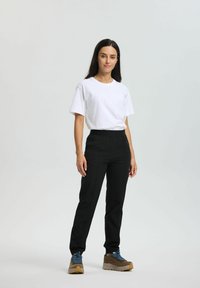 Nainen seisoo valkoisessa lyhythihaisessa paidassa, mustissa housuissa, joissa on vetoketjulliset taskut, ja ruskeissa vaelluskengissä, joissa on siniset nauhat.