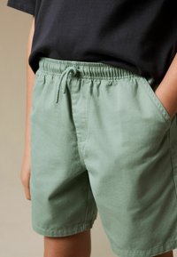 Pantaloncini verde chiaro in morbido cotone con vita elastica, coulisse e tasche laterali, caratterizzati da un design semplice e classico.