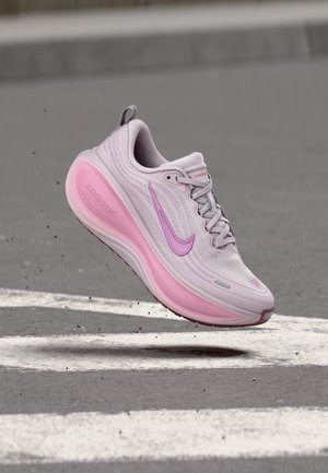 Chaussure de course Nike rose clair et violette en l’air au-dessus de l’asphalte avec des marquages routiers blancs.