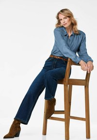 Hellblaues Denimhemd, langärmlig mit Knopfleiste; kombiniert mit dunkelblauen, geraden Jeans und braunen Wildleder-Stiefeln mit Knöchelhöhe. Sitzt auf einem Holzstuhl.