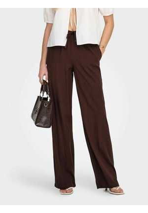 Kvinde iført løstsiddende mørkebrune bukser, hvid kortærmet top, holder en tekstureret mørkebrun håndtaske og har åbne sandaler på.