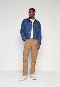 Donkerblauwe spijkerjack met twee borstzakken, gecombineerd met beige cargobroek met meerdere zakken, gedragen met witte hoge sneakers.