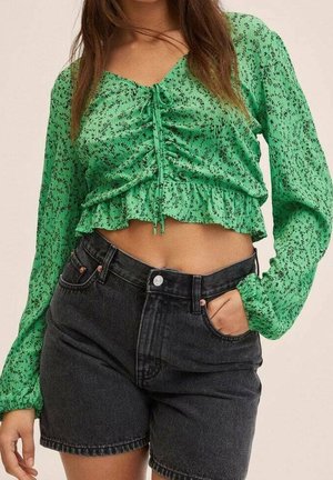 Femme portant un haut court froncé à manches longues vert et un short en jean noir taille haute, la main dans la poche.