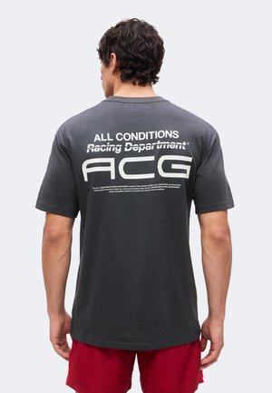 Mand med mørkt hår iført sort "ALL CONDITIONS Racing Department ACG" T-shirt og røde shorts, vist bagfra på ensfarvet baggrund.