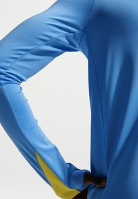 Άνω μέρος σώματος με μπλε αθλητική μπλούζα μακριάς μανσέτας με κίτρινες πατούρες στις μασχάλες, το χέρι λυγισμένο και η παλάμη ακουμπισμένη στη μέση.