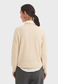 Beige knitted sweater with a textured finish and ribbed hem, worn over a collared shirt, featuring long sleeves and a relaxed fit.
