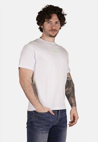 T-shirt bianco a maniche corte con un piccolo logo verde sul petto. Il modello presenta dei tatuaggi visibili sulle braccia. Materiale in cotone.