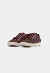 Paire de baskets basses en toile marron foncé avec semelles en caoutchouc blanches et fermeture à lacets, présentées sur un fond clair uni.