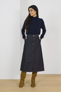 Femme debout les mains dans les poches, portant un col roulé bleu marine, une jupe midi en denim foncé et des bottes hautes en daim marron.