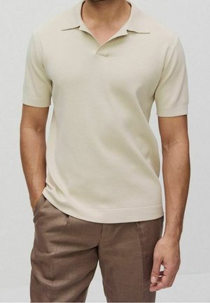 Ein Mann trägt ein beigefarbenes kurzärmeliges Poloshirt und braune Hose, mit einer Hand in der Tasche vor einem einfarbigen Hintergrund.