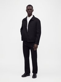 Man standing wearing a black buttoned jacket, black pants, black shoes, and a white collared shirt against a plain light background.