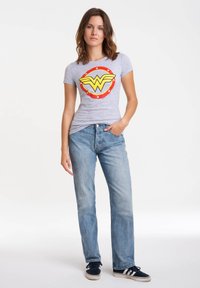 Gray cotton t-shirt with a red, yellow, and blue Wonder Woman logo, paired with light blue jeans and dark sneakers.