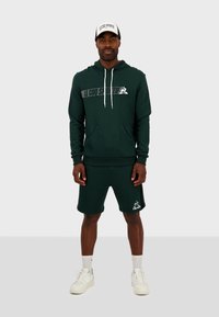 Sudadera de algodón verde con bolsillo frontal y logotipo, combinada con pantalones cortos a juego. Zapatillas blancas y una gorra en blanco y negro completan el look.